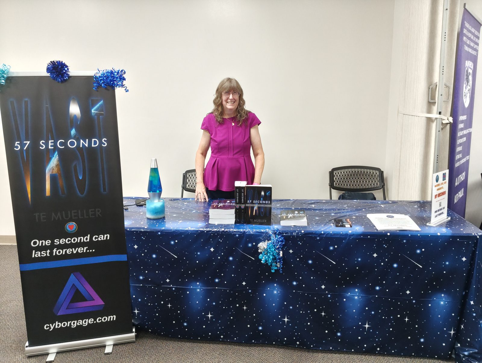 Teri Mueller behind the CyborgAge booth table at NerdyCon 2026, with a Vast: 57 Seconds banner and copies of the book on a starfield-print tablecloth